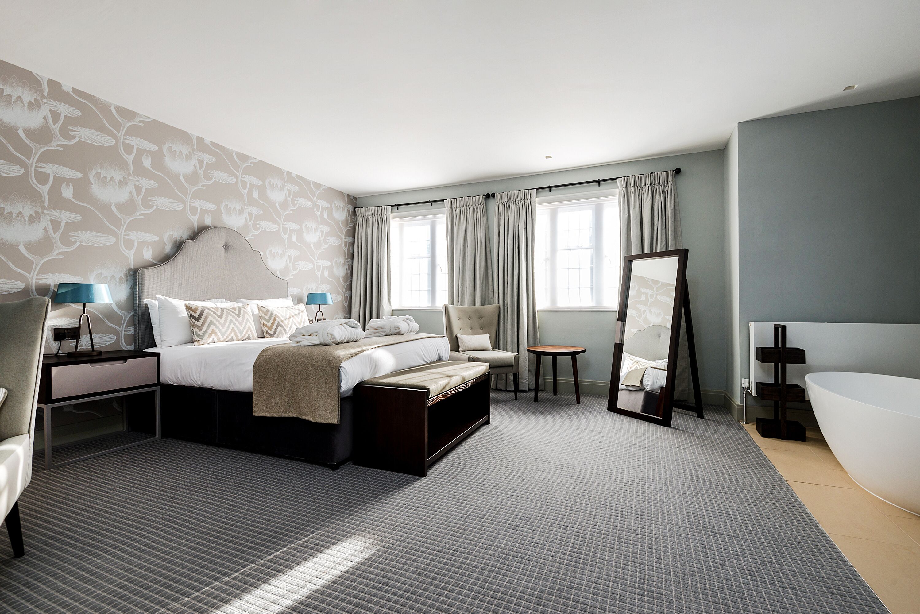 Una habitación de hotel moderna con una cama grande, una zona de estar, una mesita, un espejo y una bañera.