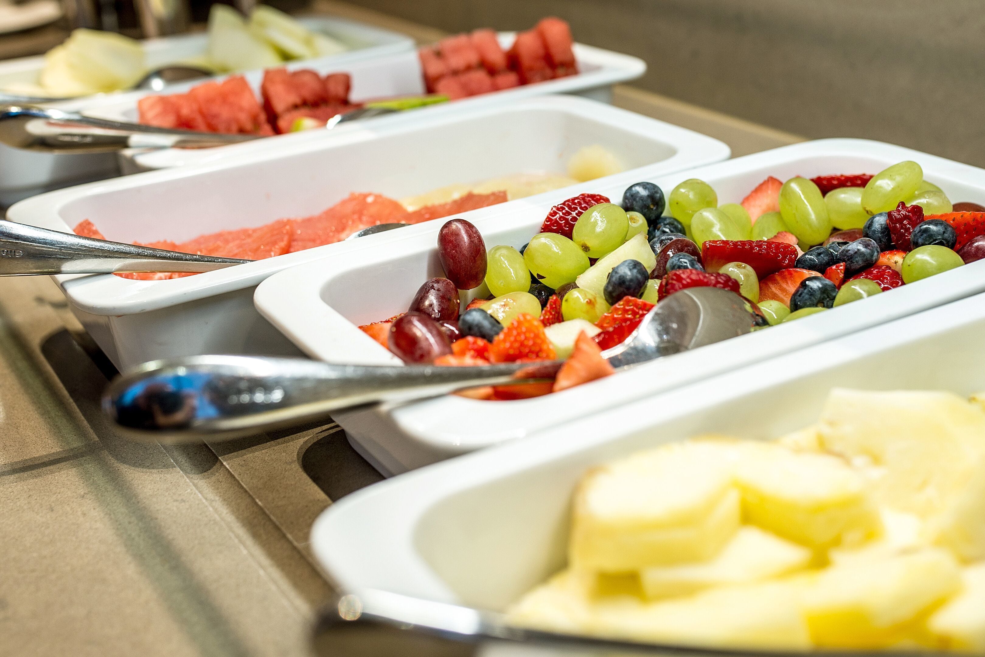A buffet with various fruits and vegetables in white trays, including watermelon, grapes, strawberries, and green beans.