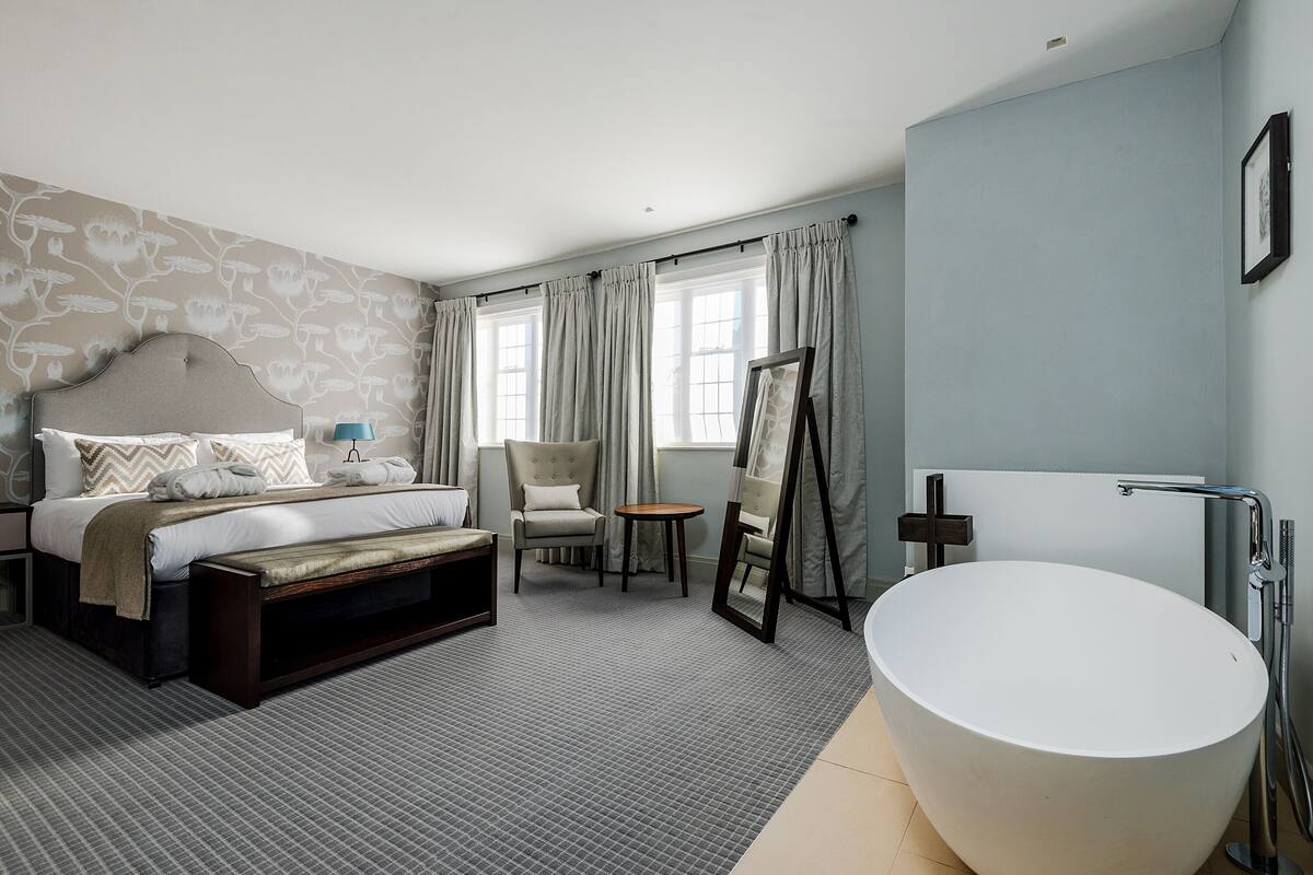 Una habitación de hotel moderna con una cama grande, un área de descanso con una silla y una mesa, una bañera amplia y una ventana con cortinas.