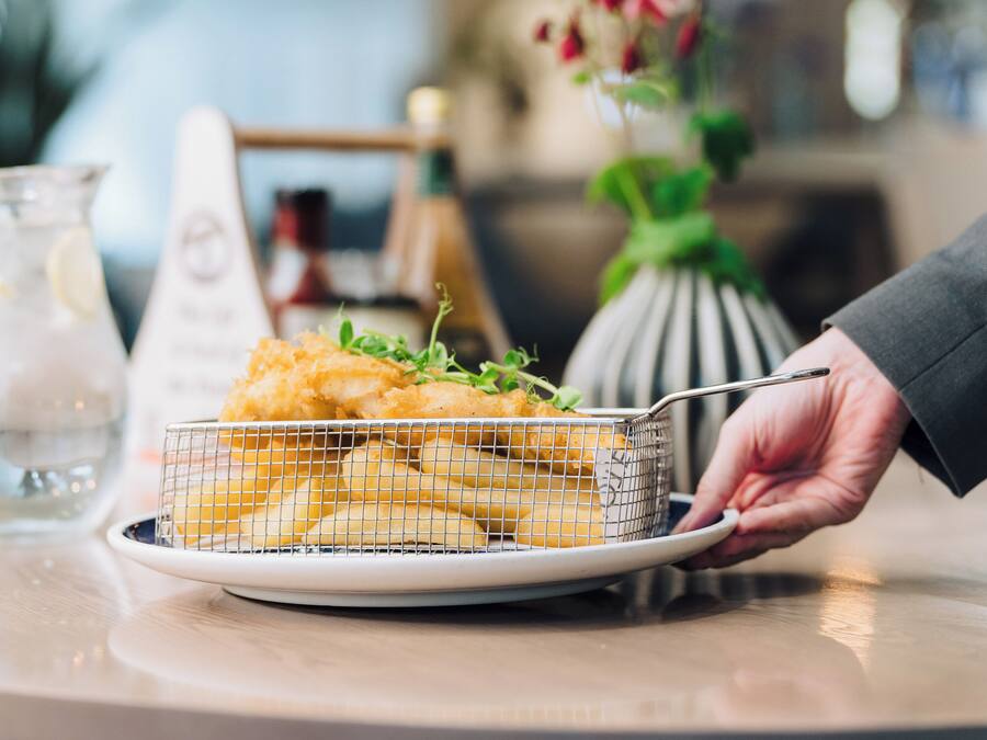 Une main tenant un panier rempli de fritures sur une assiette, avec un pichet et un vase en arrière-plan.