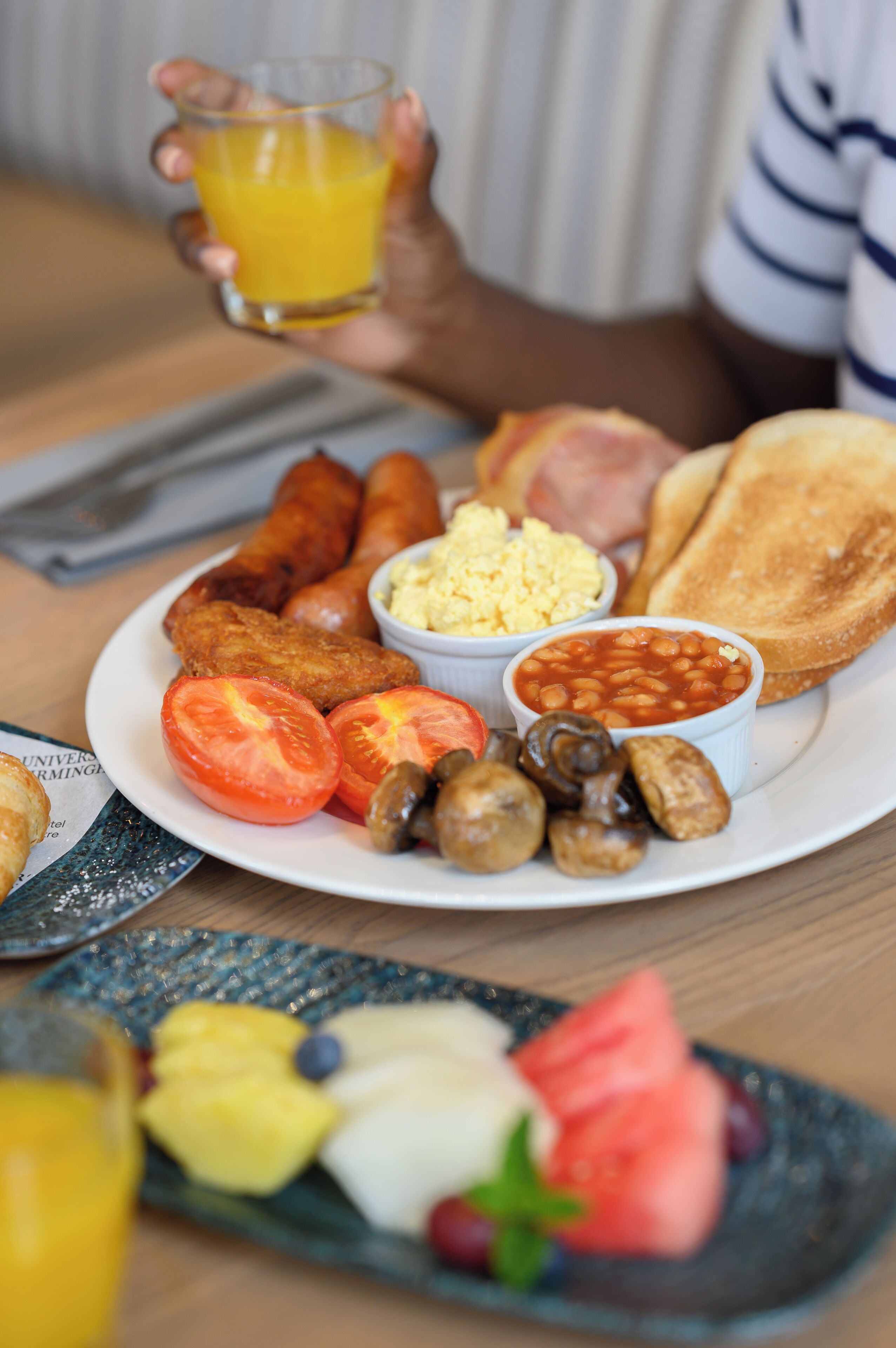 Une assiette de petit-déjeuner comprenant des œufs, des saucisses, des toasts, des tomates, des champignons, des haricots et un verre de jus d’orange.