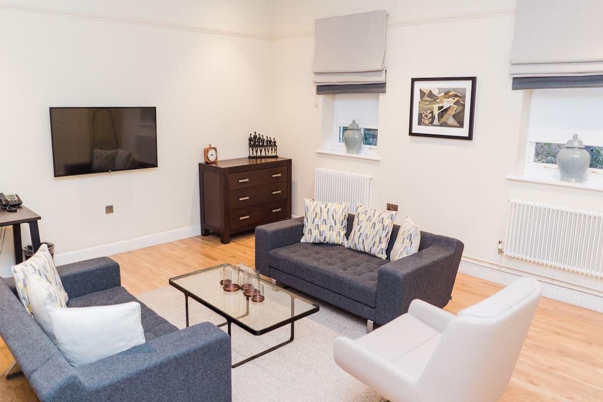 Una sala de estar moderna con un sofá gris, un sillón blanco y una mesa de centro de vidrio. Hay un aparador de madera oscura, un televisor de pantalla plana y una ventana con vistas.
