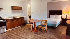 A compact hotel room with a kitchenette, a dining area with two chairs, a bed, and a nightstand with a lamp. - Jefferson Inn and Suites (Newport News)