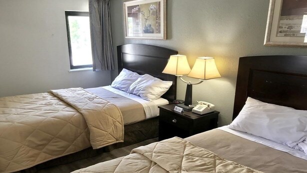 A hotel room with two beds, each with a quilted bedspread, a nightstand with a lamp, and a framed picture on the wall. - Jefferson Inn and Suites (Newport News)