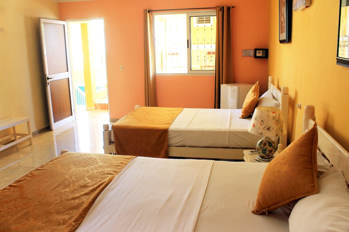 A hotel room with two beds, a window with curtains, a bedside table with a lamp, and a small refrigerator.