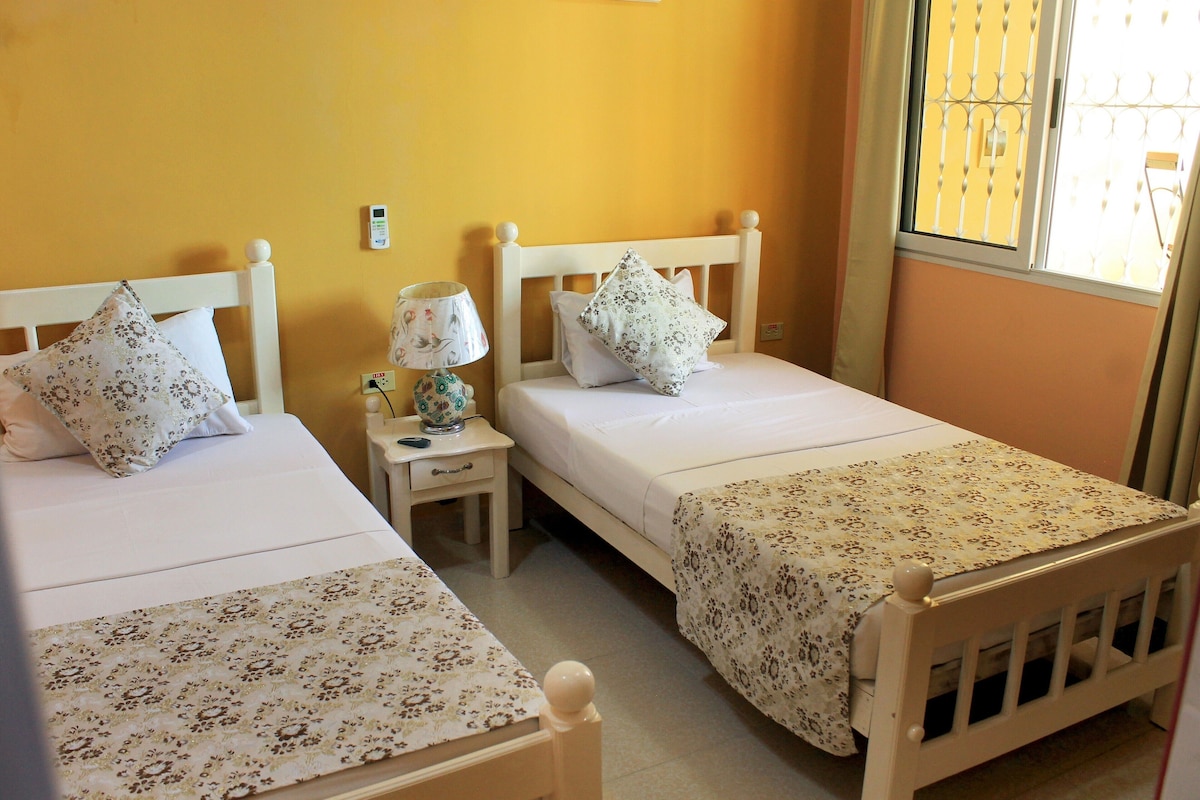 A bedroom with two single beds, a bedside table with a lamp, and a window with a metal grill.