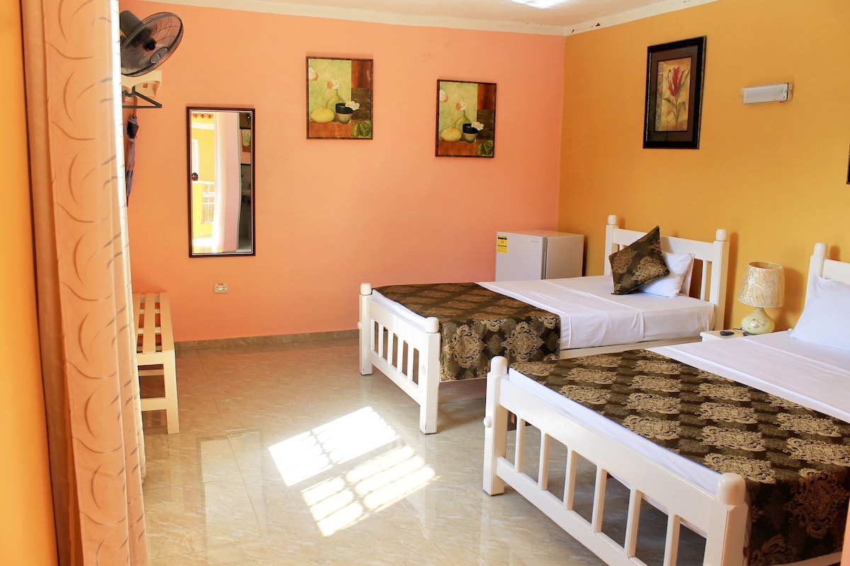 A room with two beds, a small table, and a chair. There are two wall-mounted fans and three framed pictures on the wall.