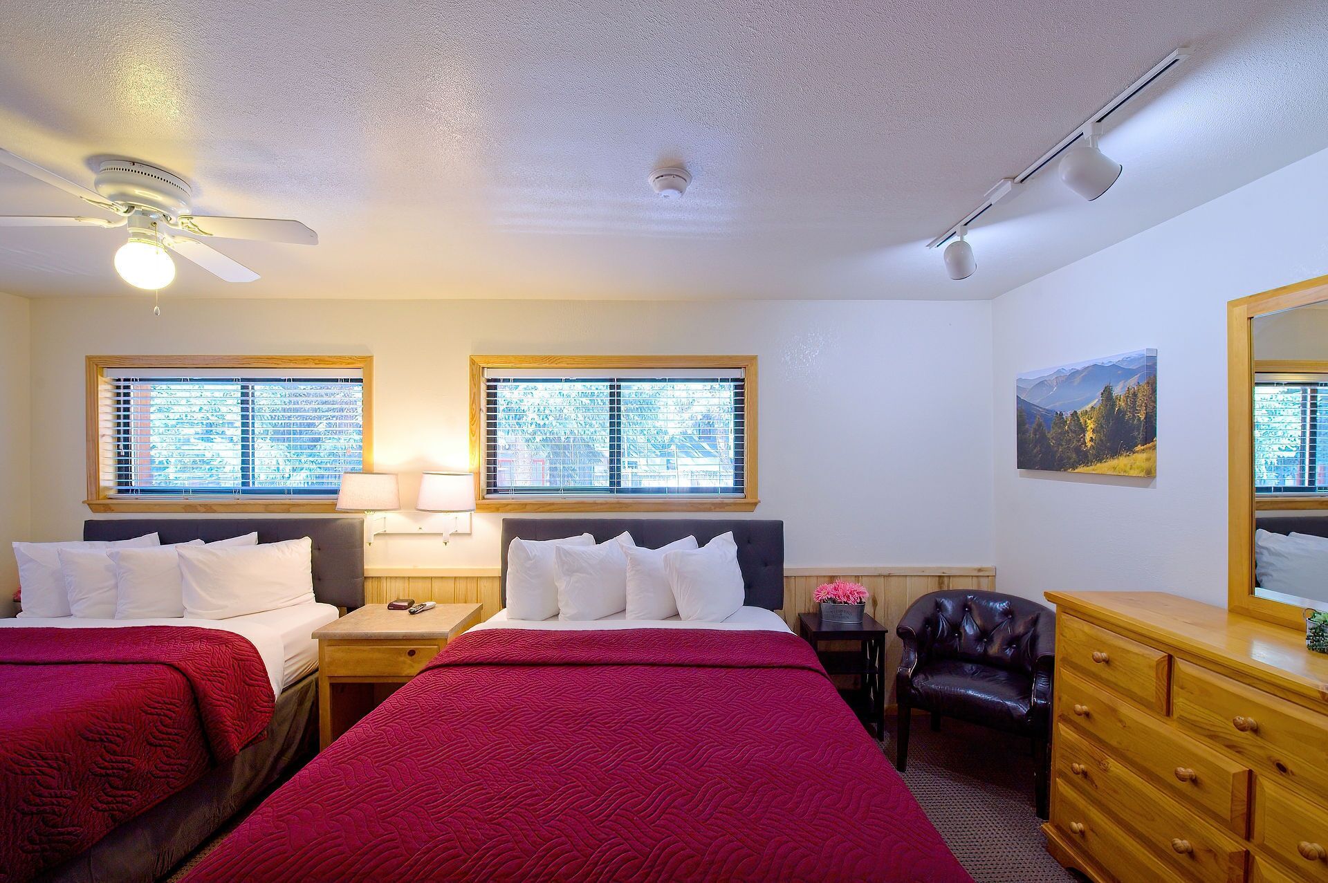 Une chambre d’hôtel avec deux lits, un ventilateur de plafond, une fenêtre avec des stores, un bureau avec une lampe, une chaise, une commode et un tableau au mur.