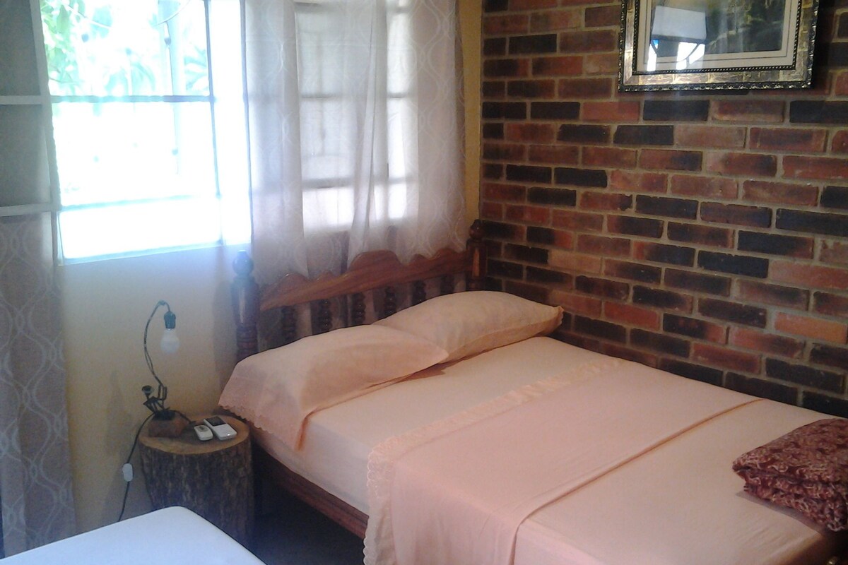 A single bed with a wooden headboard, a bedside table with a lamp, and a brick wall with a framed picture.