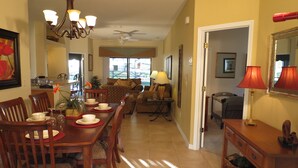 A spacious dining area with a long wooden table set for four, flanked by wooden chairs. A chandelier hangs above the table, and a ceiling fan is visible in the background.