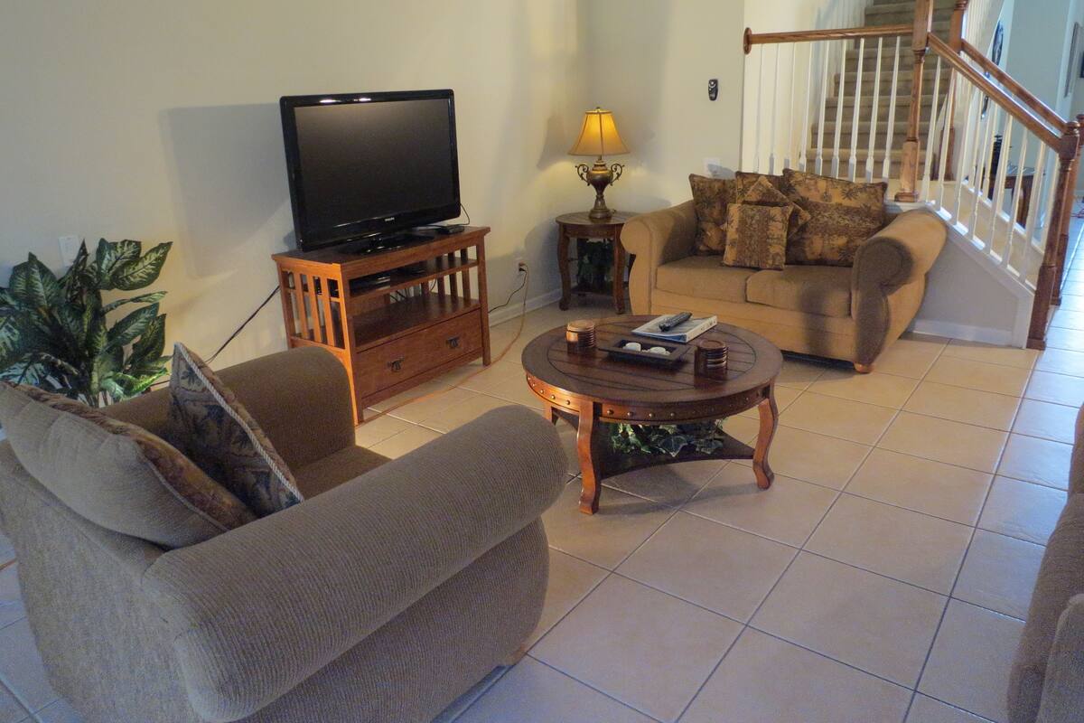 Un salon avec un téléviseur posé sur un meuble en bois, un canapé, une table basse et un escalier en arrière-plan.