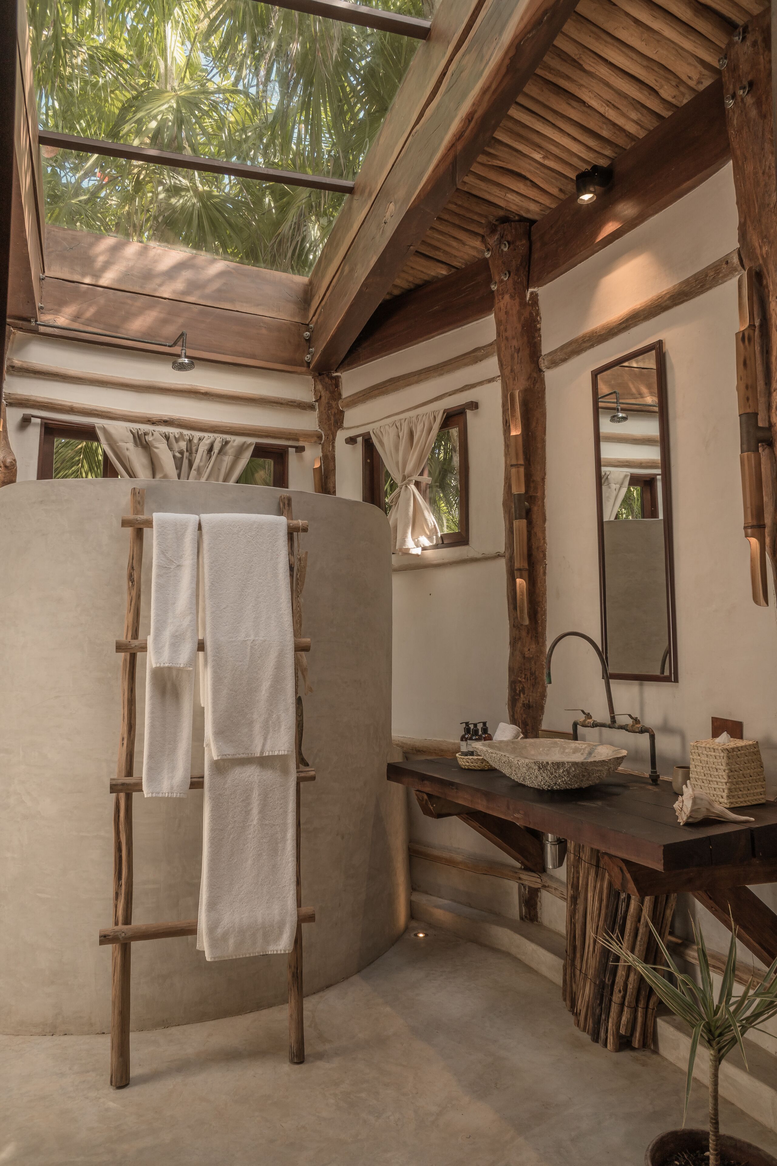 Un baño con una escalera de madera, un lavabo, un espejo y vistas a palmeras a través de un tragaluz.