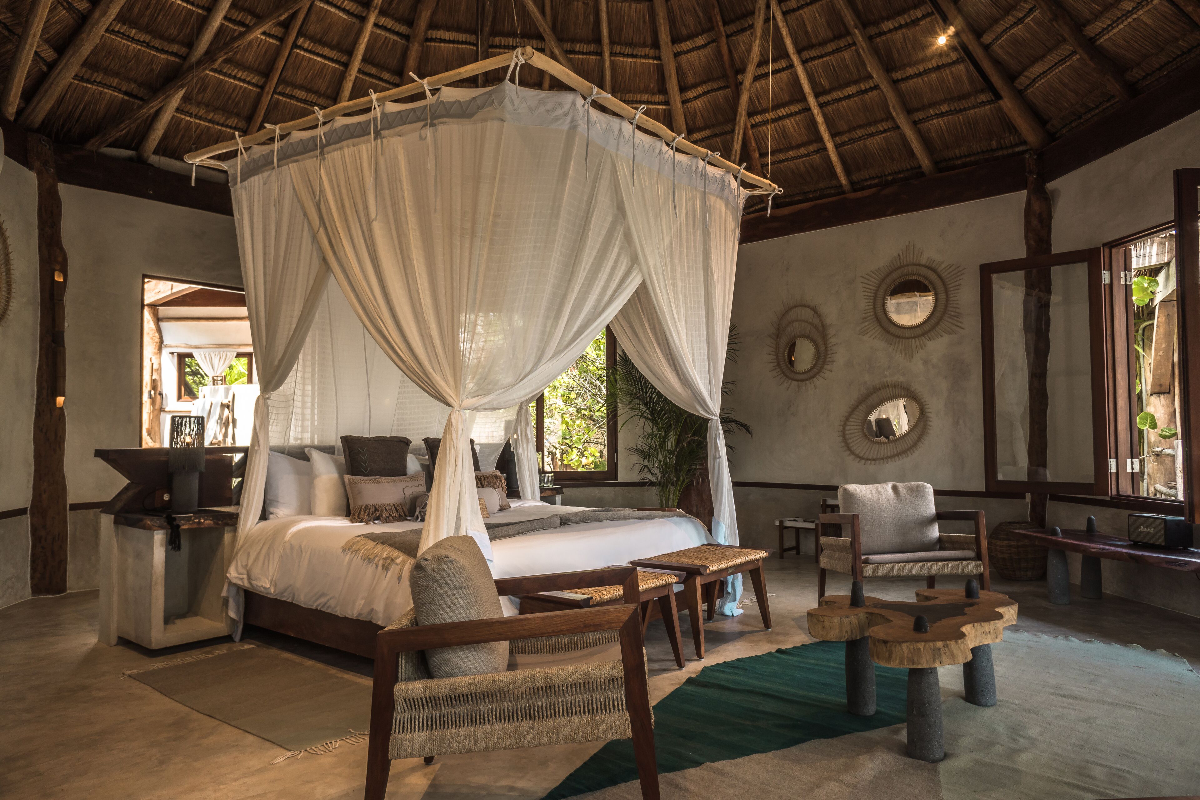 A spacious bedroom with a four-poster bed, a sitting area with a chair and table, and a large window providing natural light.