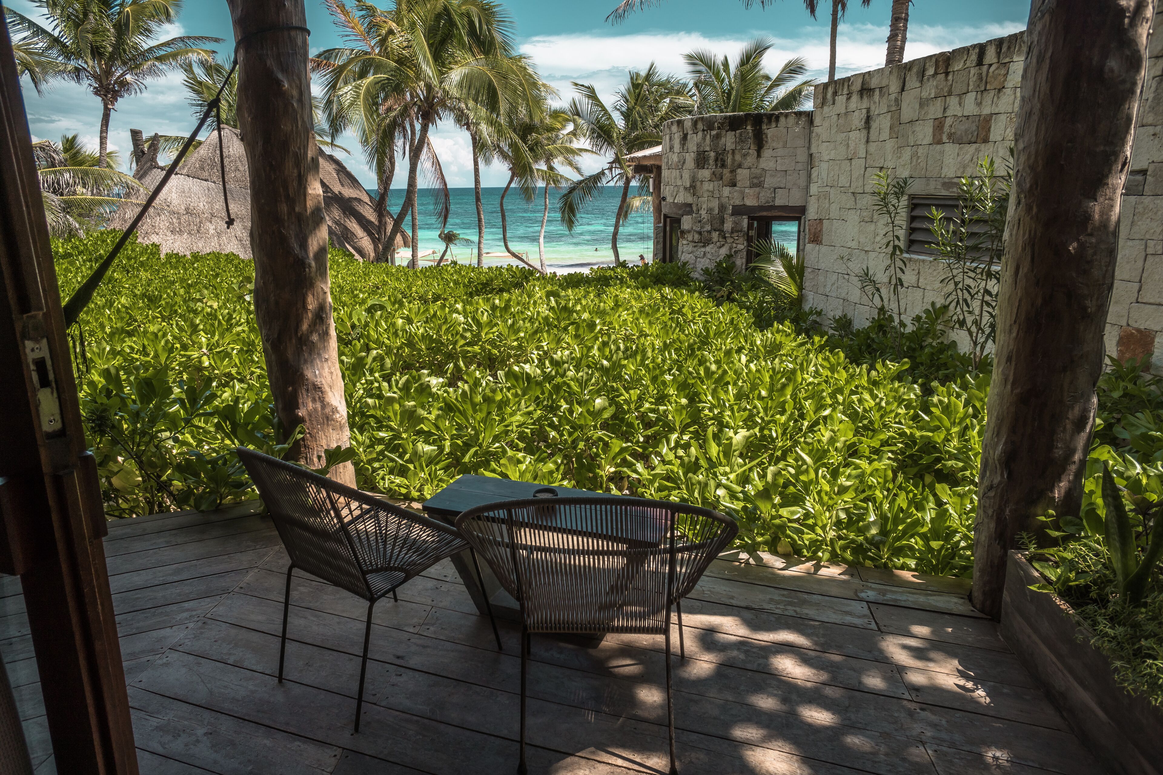 Une terrasse en bois avec deux chaises et une table, donnant sur un jardin luxuriant et verdoyant et sur une plage bordée de palmiers.