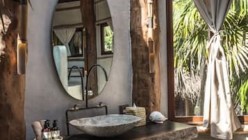 A rustic bathroom with a stone sink, wooden beams, a large mirror, and a view of palm trees.
