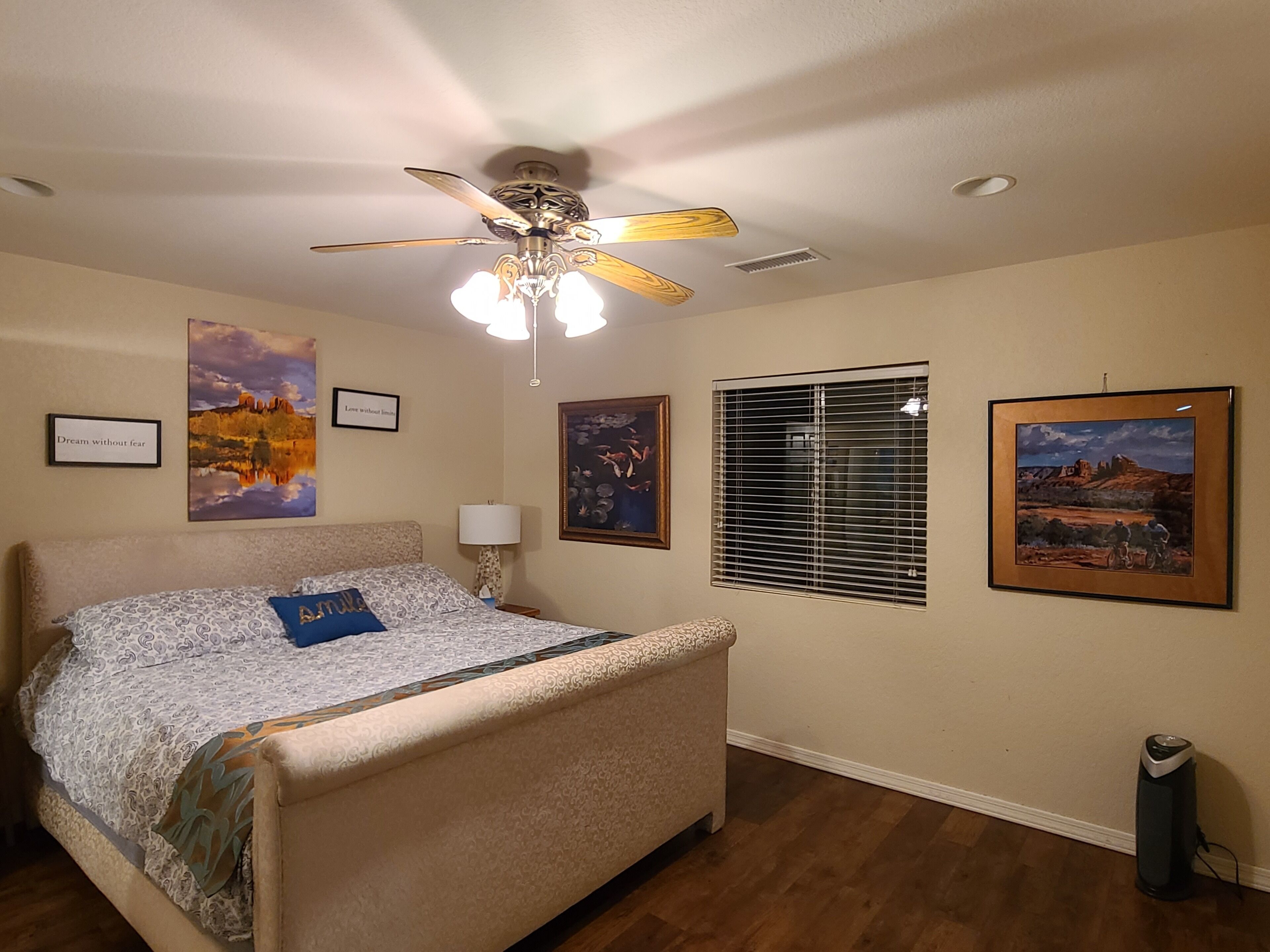 A bedroom with a bed, ceiling fan, wall art, and a window with blinds.