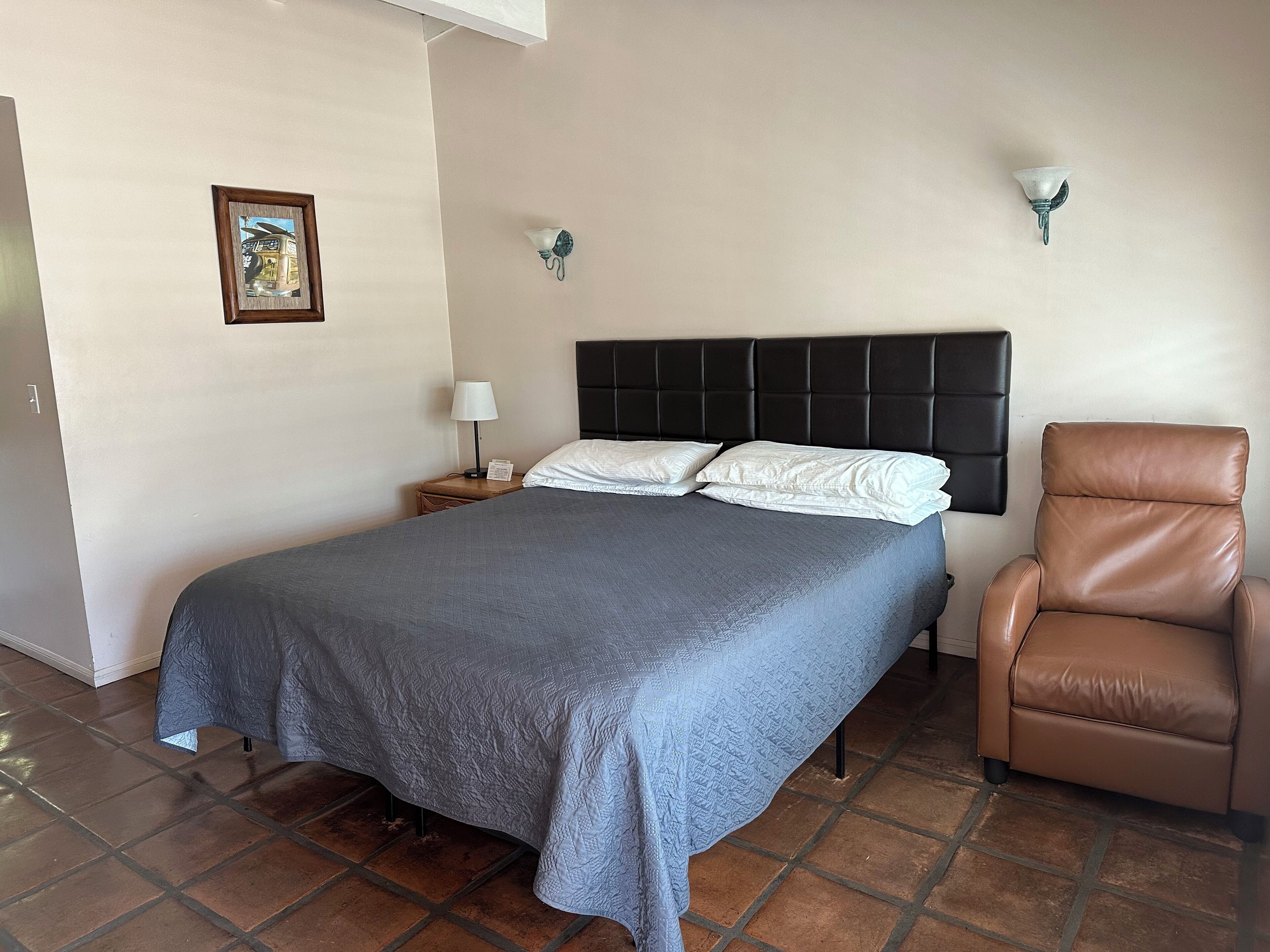 A neatly made bed with a grey quilt, a brown leather chair, a small bedside table with a lamp, and two wall-mounted light fixtures.