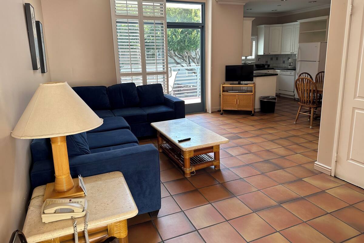 A living room with a sofa, a small table, a lamp, and a telephone. There is a kitchen area visible through an open door.