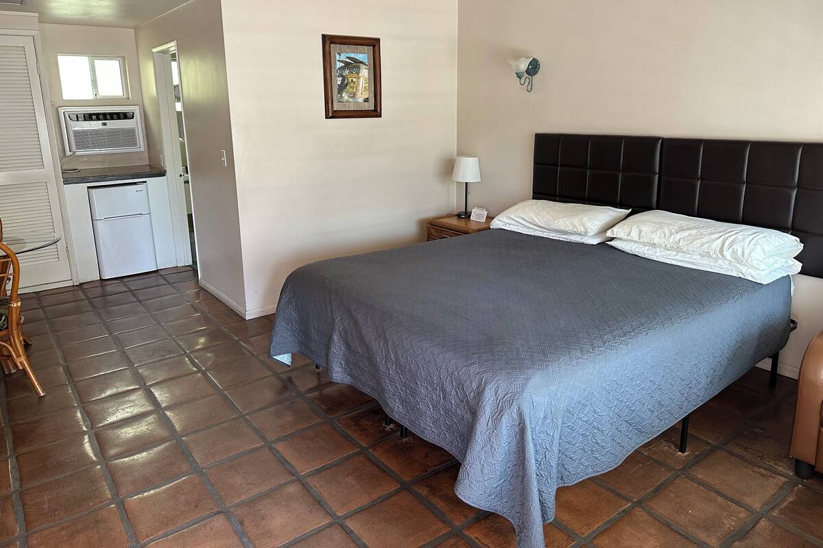 A bedroom with a bed, a nightstand, a lamp, a picture on the wall, and a kitchenette area with a refrigerator and an air conditioner.