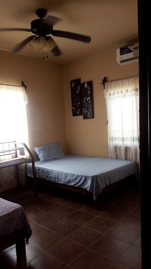 Una habitación pequeña con cama individual, ventilador de techo, ventana con cortinas traslúcidas y aire acondicionado de pared.