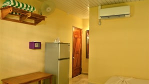 Quarto compacto com um banco de madeira, geladeira, ventilador e porta que dá para outro cômodo.