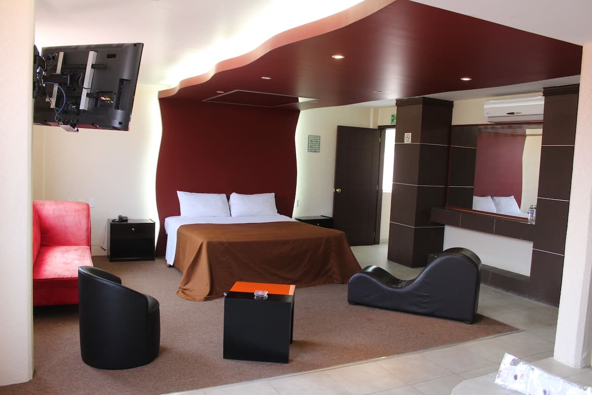 A hotel room with a bed, a red chair, a black ottoman, a small table, and a large mirror.