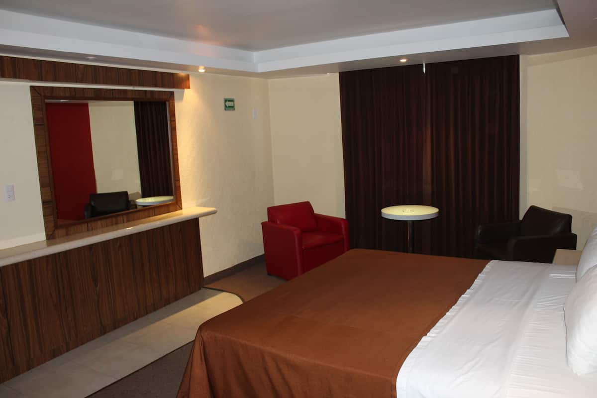 A hotel room with a large bed, a red chair, a small round table, and a mirror above the counter.