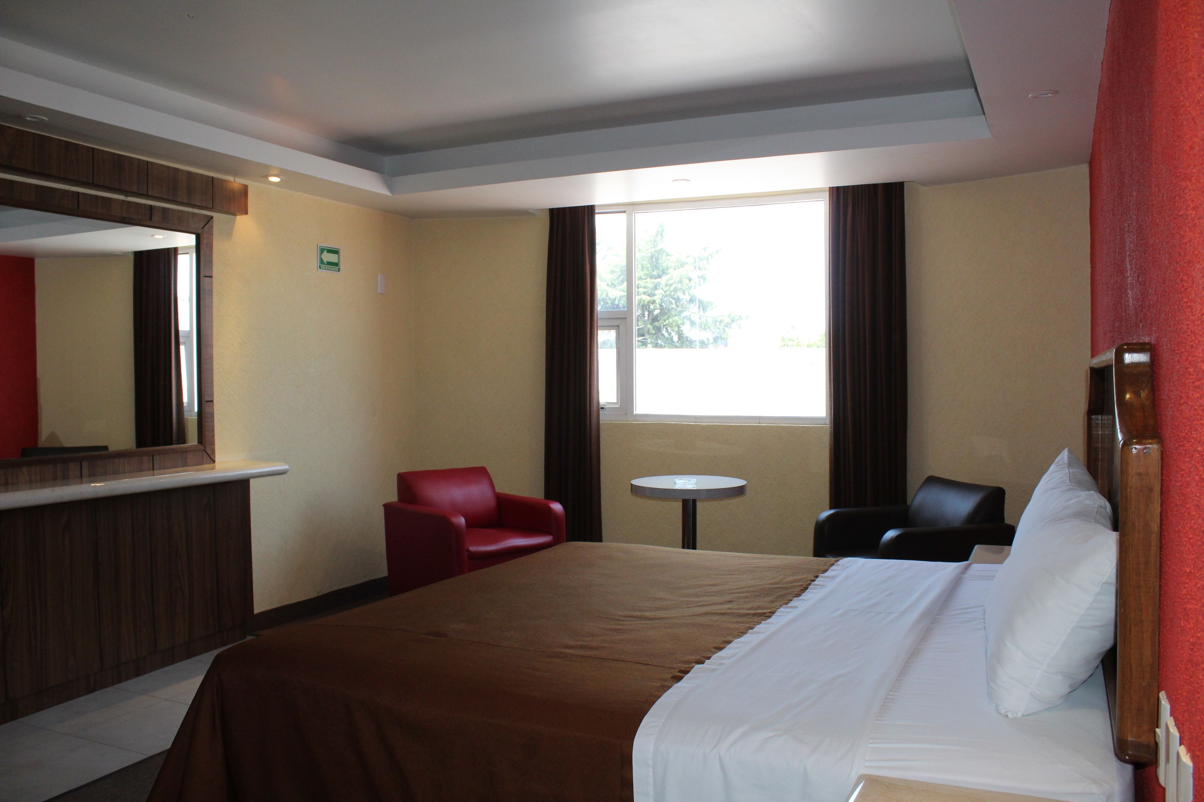 A hotel room with a large bed, a red armchair, a small round table, and a window with curtains.