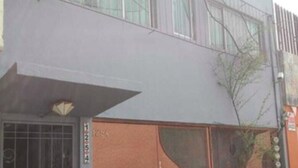 Un edificio con un amplio portón de garaje, una entrada protegida por una reja y un pequeño árbol en primer plano.