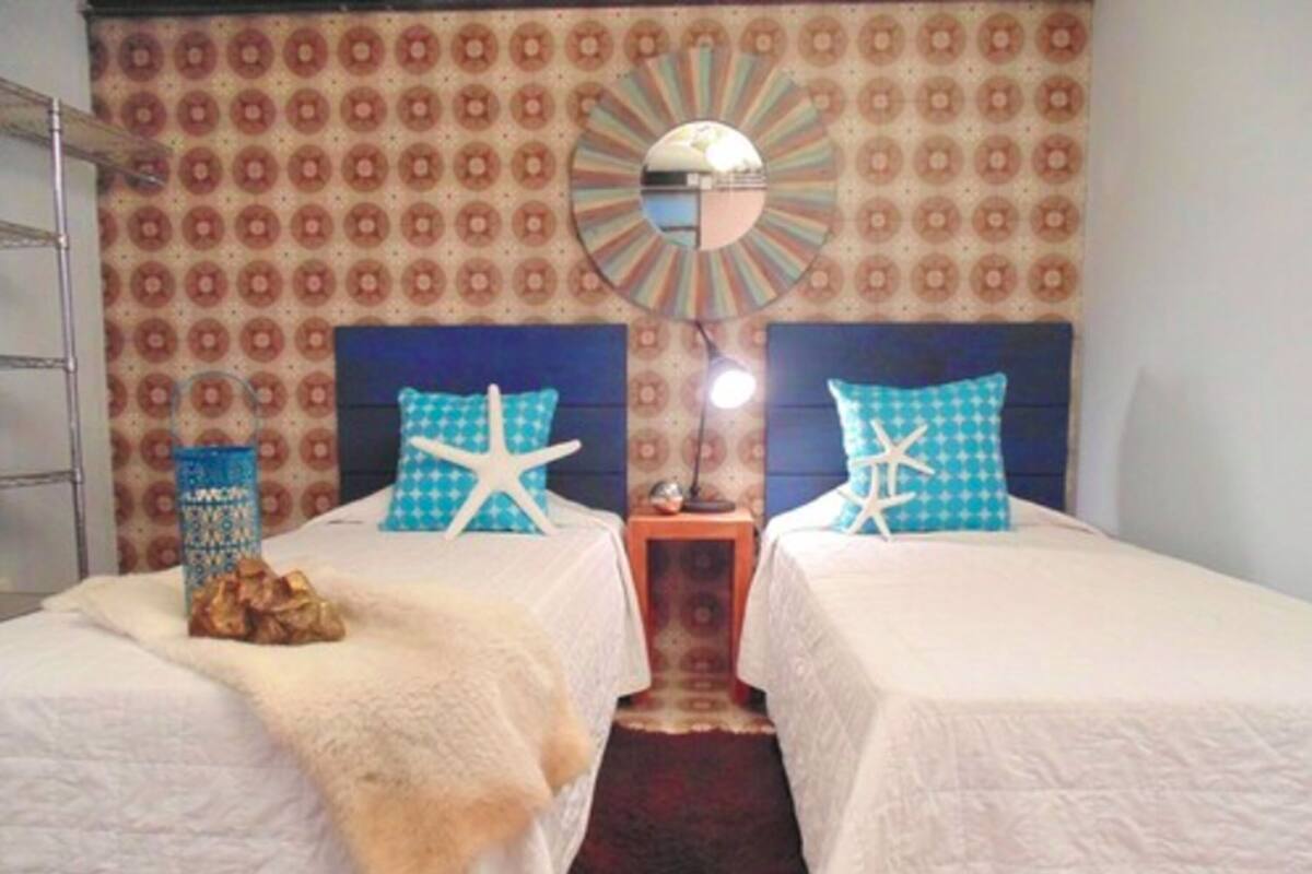 Une chambre avec deux lits, chacun recouvert d’une courtepointe blanche et agrémenté d’oreillers en forme d’étoile de mer, un miroir au-dessus du lit et un papier peint à motifs.