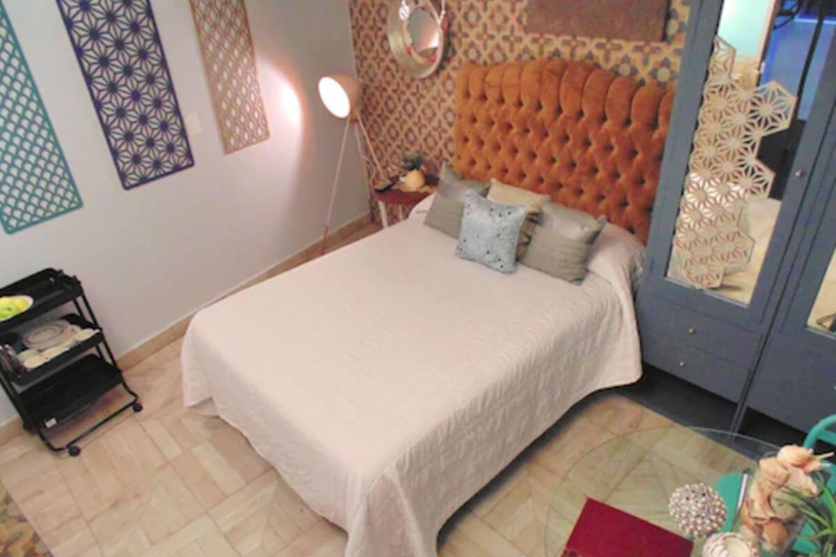Un lit bien fait, recouvert d’une courtepointe blanche et agrémenté de plusieurs oreillers, une petite table de chevet avec une lampe et un panneau décoratif au mur.