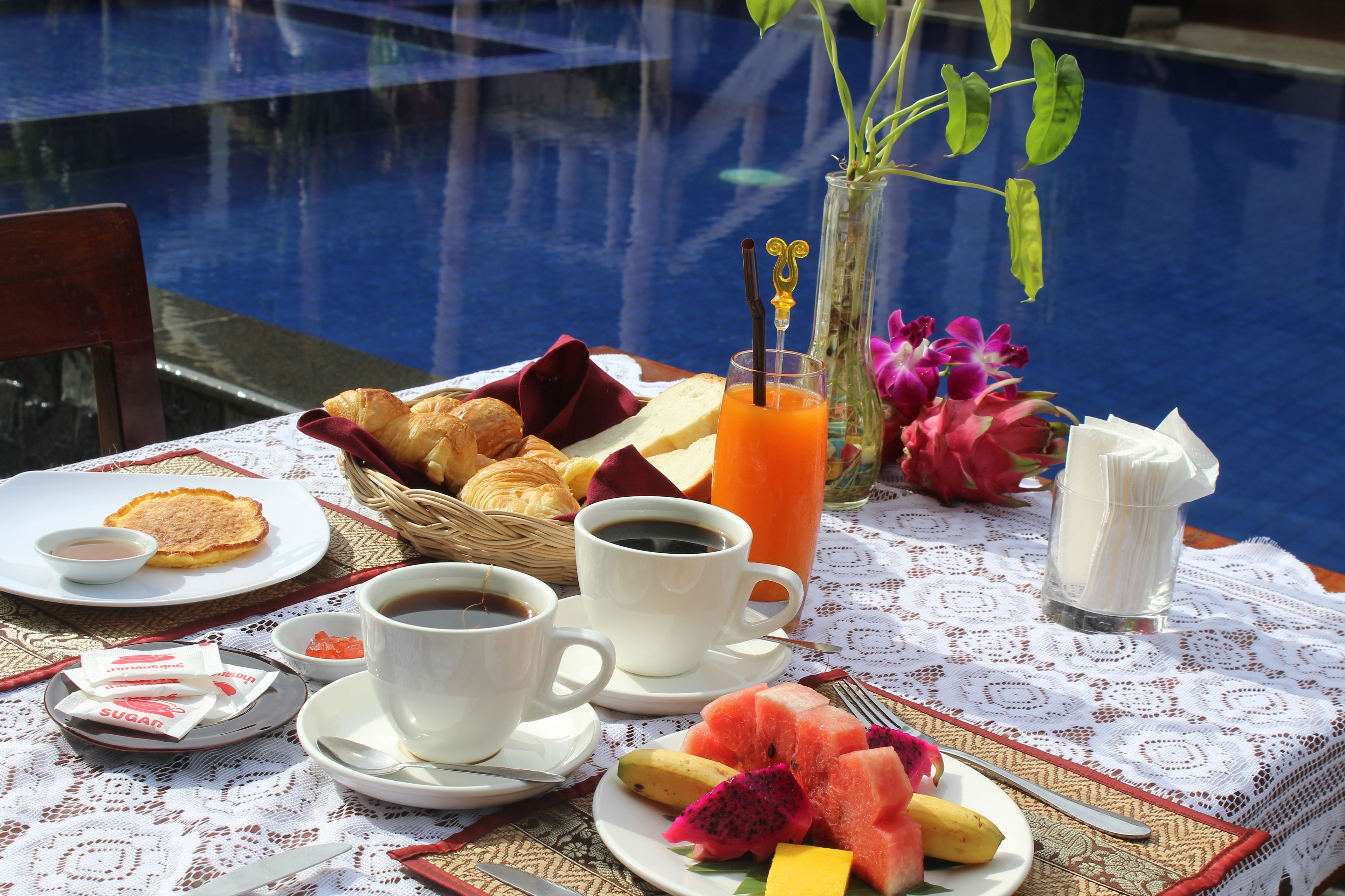 A breakfast setup by a pool with croissants, fruit, coffee, and juice.