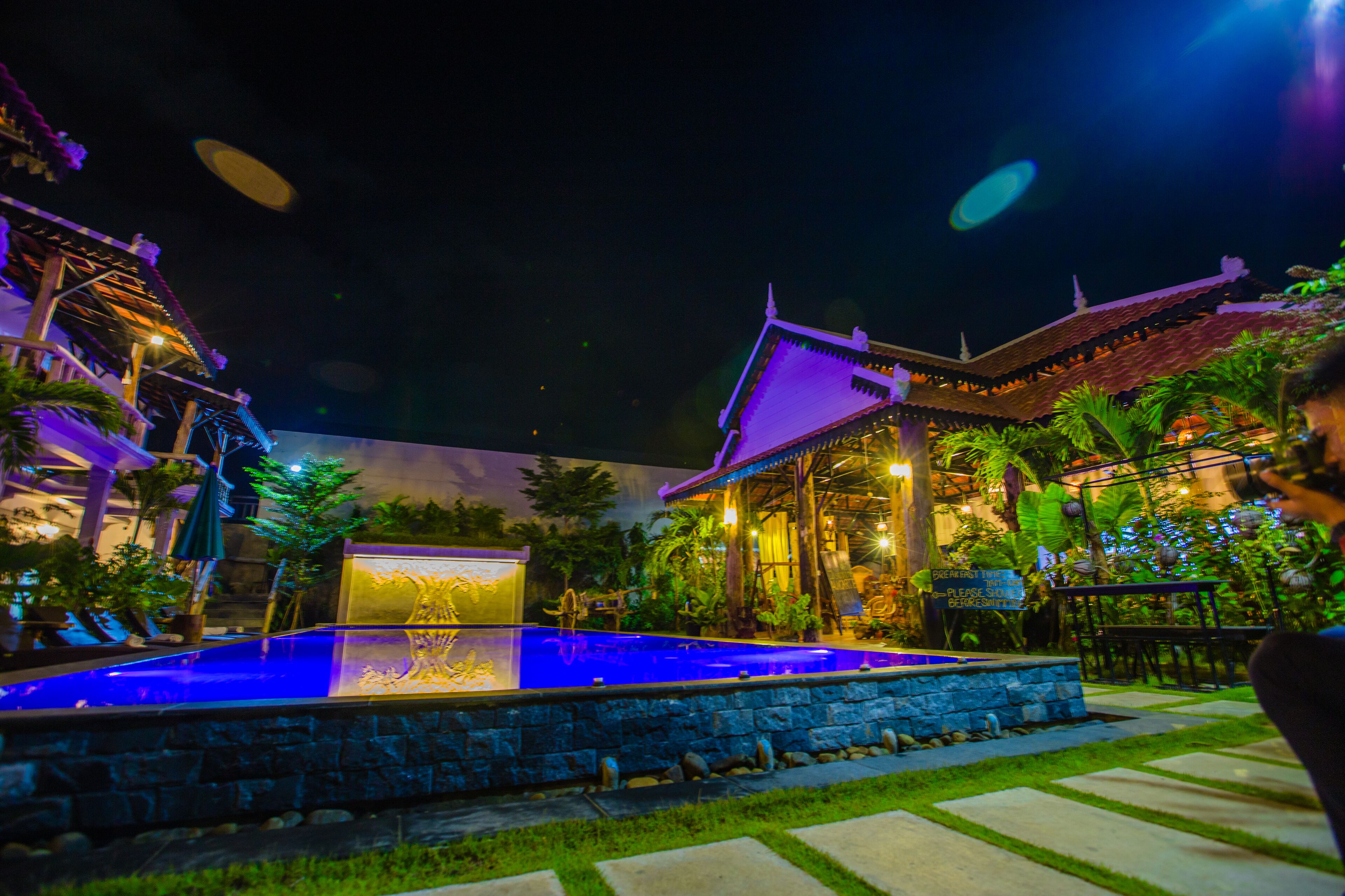 Vista nocturna de un patio con piscina, arquitectura tradicional y abundante vegetación.