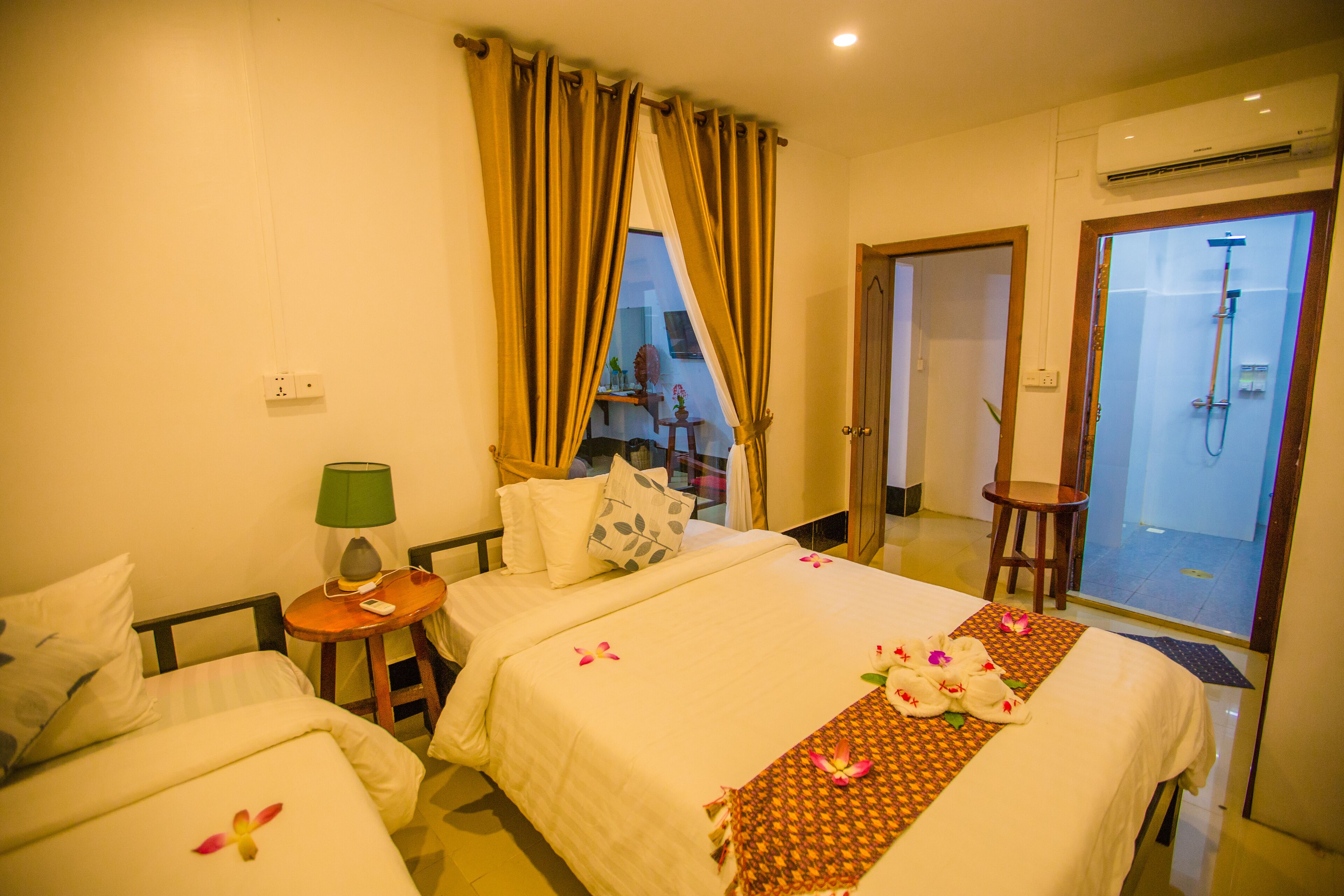 A hotel room with two beds, a small table with a lamp, a shower area, and a view of the outside.