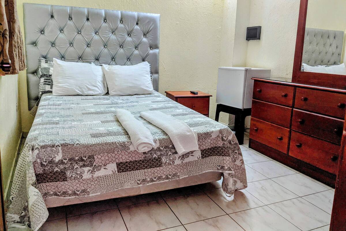 Una cama bien hecha con una colcha estampada y almohadas, un aparador de madera con varios cajones, y una mesita de noche con una lámpara.