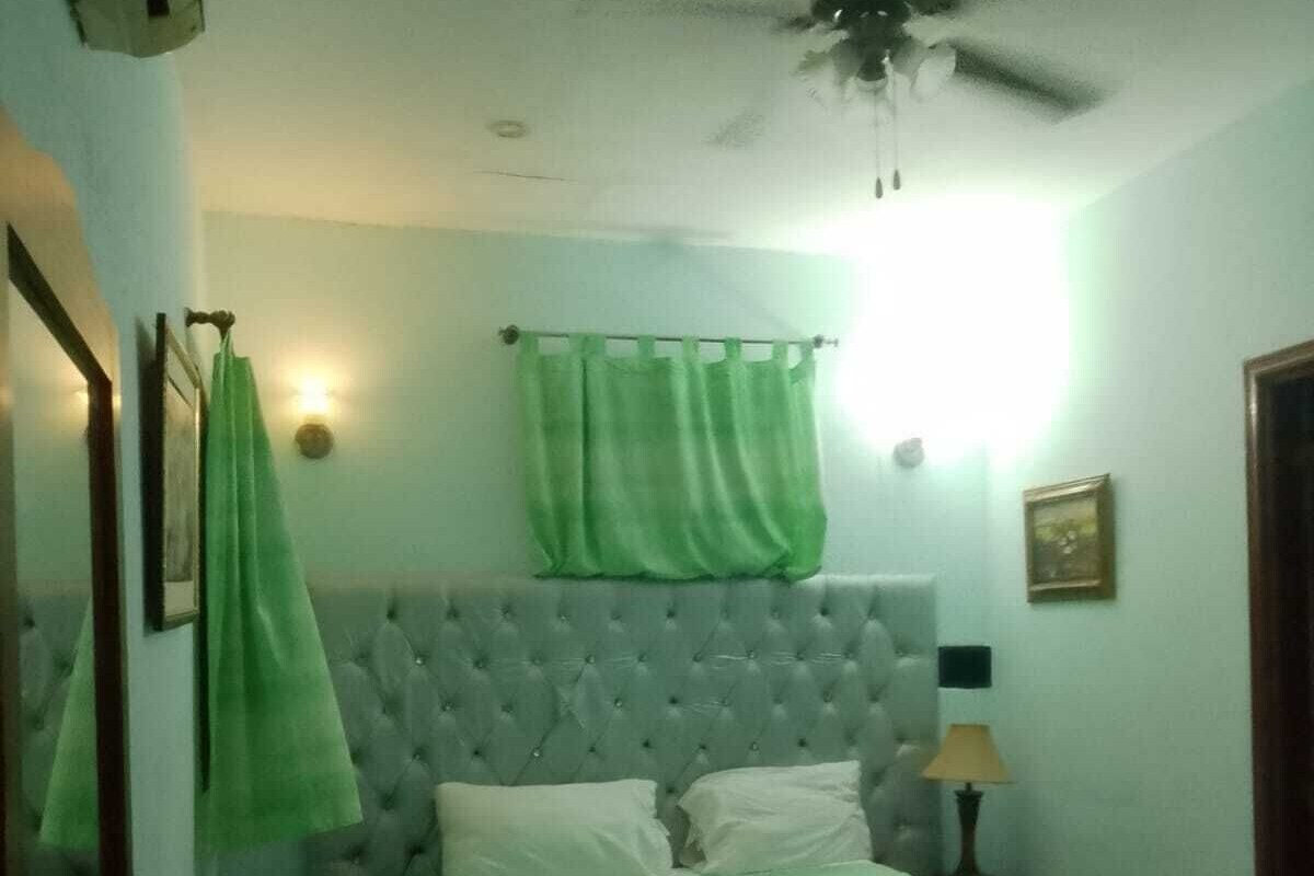 Een slaapkamer met een gestoffeerd hoofdeinde, een bed met een turquoise en blauwe dekbedovertrek, een plafondventilator en een nachtkastje met een lamp.