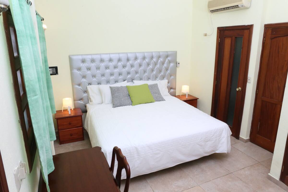 Un dormitorio ordenado con cabecera capitonada, cama con ropa blanca y un almohadón verde, dos mesitas de noche con lámparas, y una silla de madera.