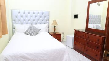 Un dormitorio ordenado con una cama grande, un aparador de madera, una lámpara de noche y un pequeño electrodoméstico blanco.