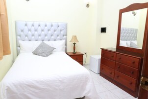 Un dormitorio ordenado con una cama grande, un aparador de madera, una lámpara de noche y un pequeño electrodoméstico blanco.
