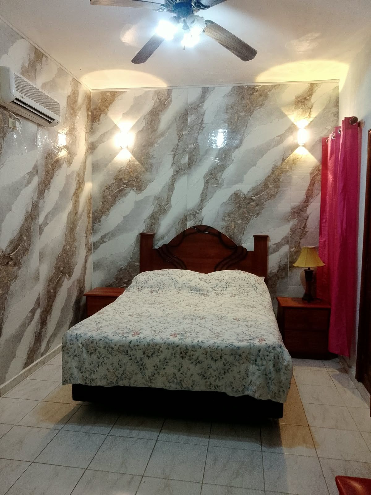 Un dormitorio con cama, ventilador de techo, aire acondicionado de pared y mesita de noche con lámpara.