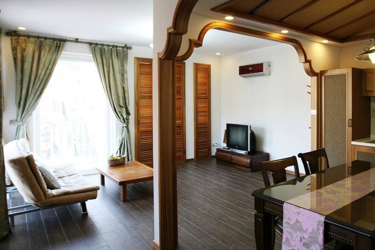 A room with a dining table, chairs, a sofa, a television, and a kitchen area with wooden cabinets and a sink.