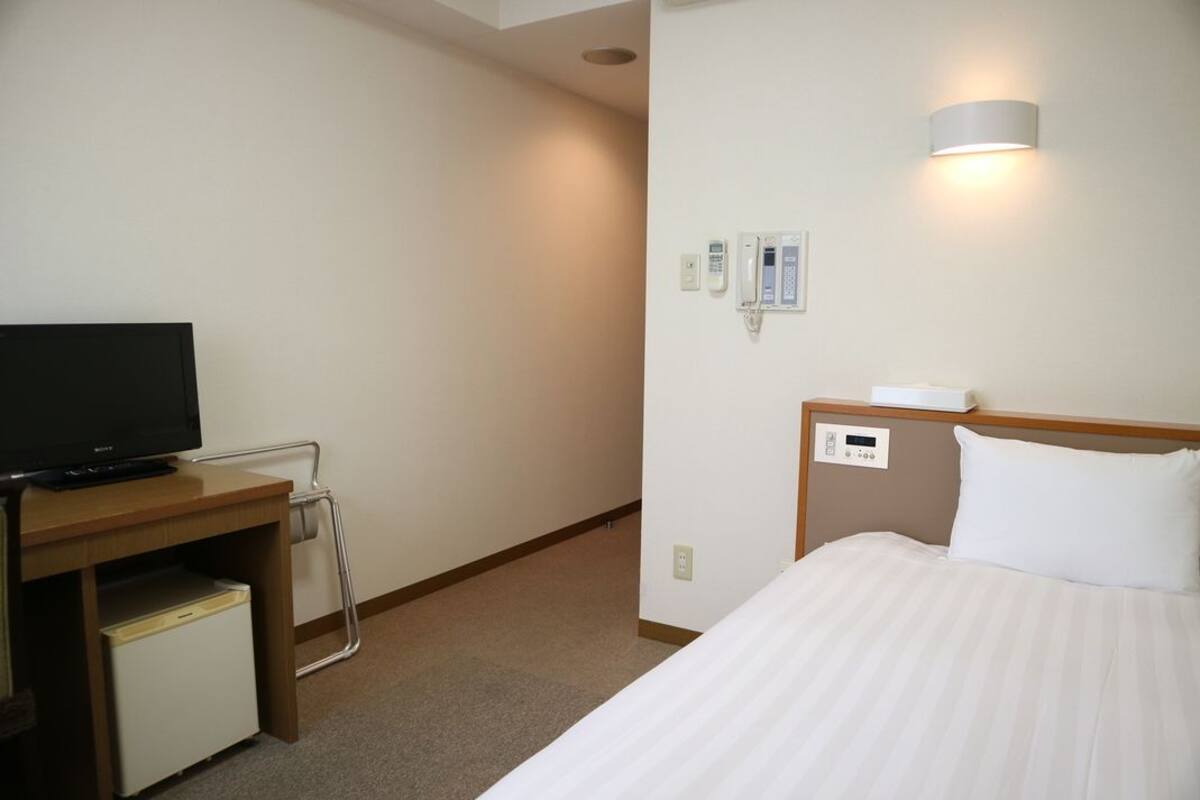 A hotel room with a bed, a TV on a desk, a bedside table, a lamp, and a wall-mounted phone.