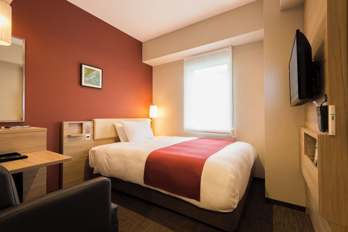 A hotel room with a bed, bedside table, lamp, TV, and a window.