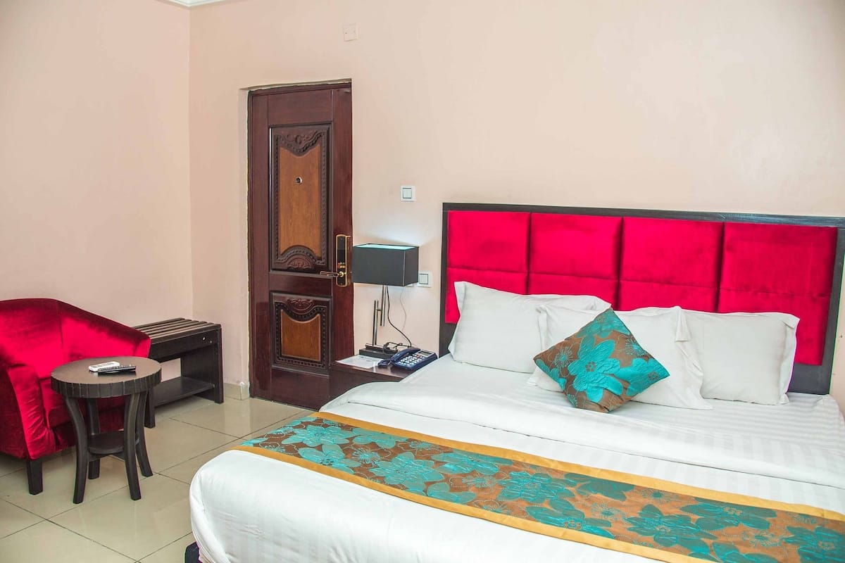Una habitación de hotel con una cama, una silla roja, una mesita y una mesita de noche con lámpara.