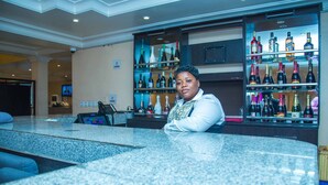 A bar area with a granite countertop, a woman seated behind the bar, and shelves stocked with various bottles of liquor. - Royal Crest Hotel & Suites (Port Harcourt)