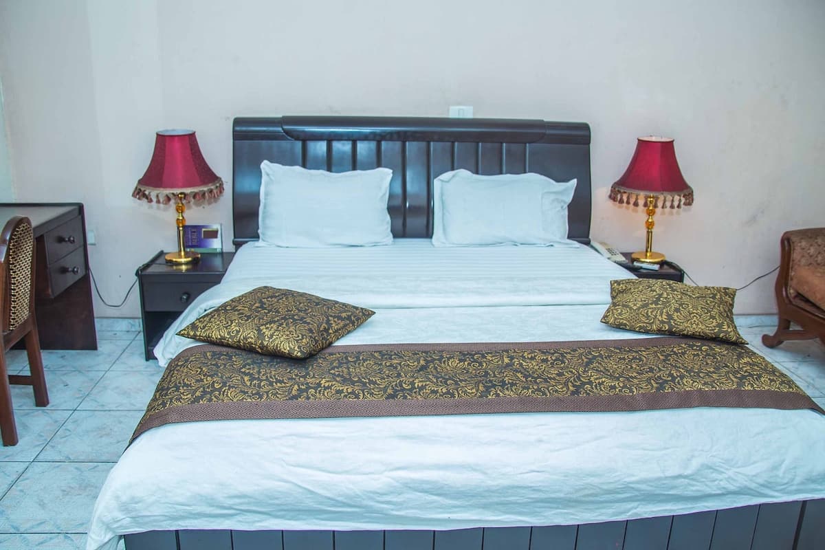Una cama bien hecha con ropa de cama blanca y dorada, dos lámparas de noche y una mesita de noche de madera con un reloj.