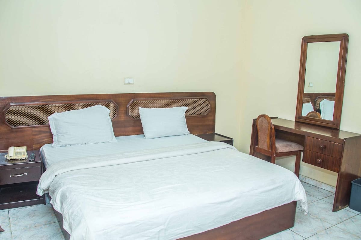 Un dormitorio con una cama de madera, dos almohadas, una mesita de noche con un teléfono, un tocador con espejo y una silla.