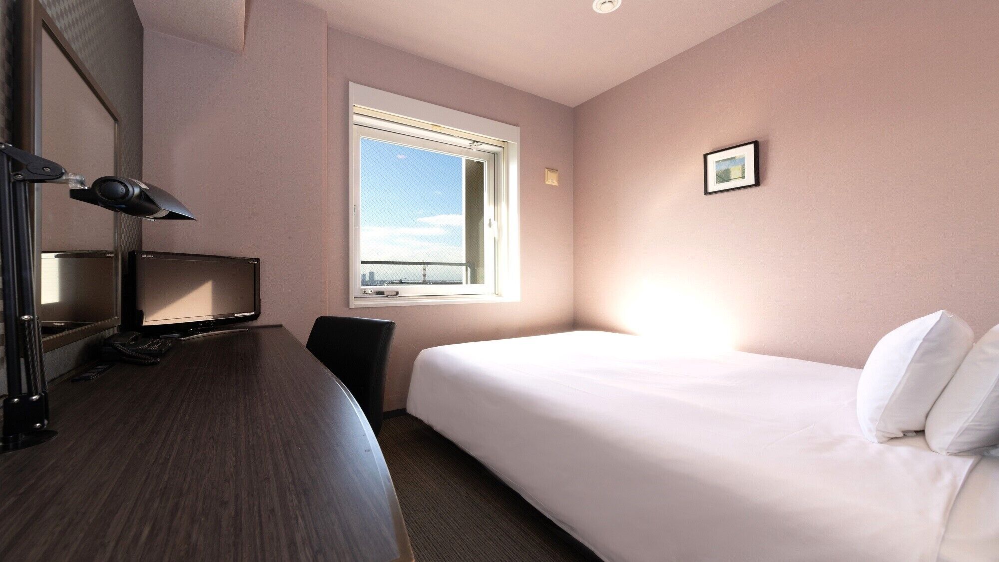 A hotel room with a bed, a desk with a computer, a chair, a window, and a picture on the wall.