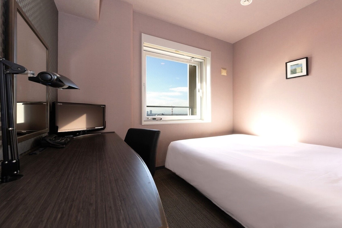 A hotel room with a bed, a desk with a computer, a chair, a window, and a picture on the wall.
