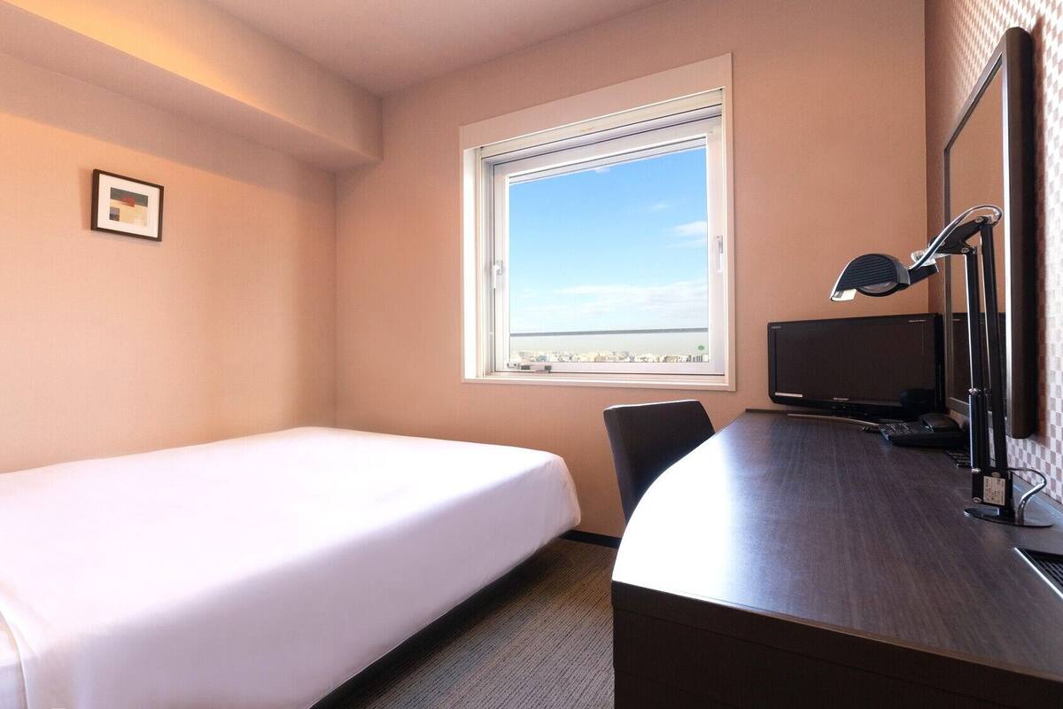 A hotel room with a bed, a desk with a computer, a chair, a window with a city view, and a framed picture on the wall.